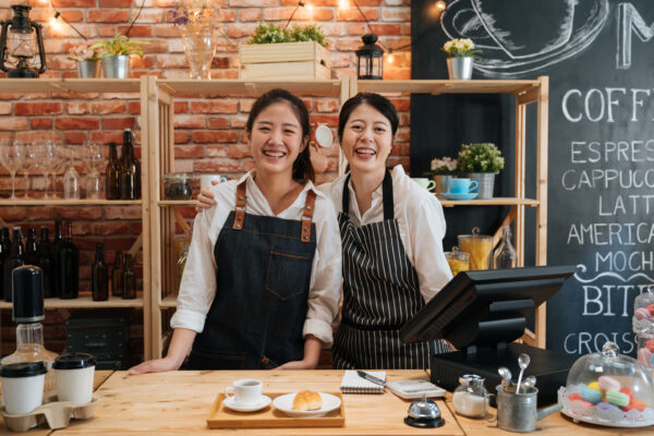 Small startup business owner concept. two successful young baristas women standing in bar counter in café. happy coffeehouse waitresses in aprons smiling confidently to camera in coffee shop.