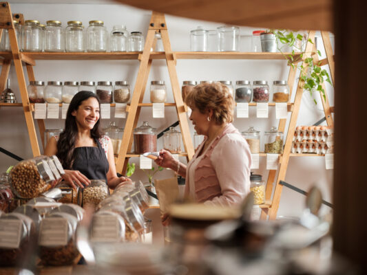 Happy hispanic employee and senior female customer standing next to the shelves of a sustainable shop and smiling at each other.