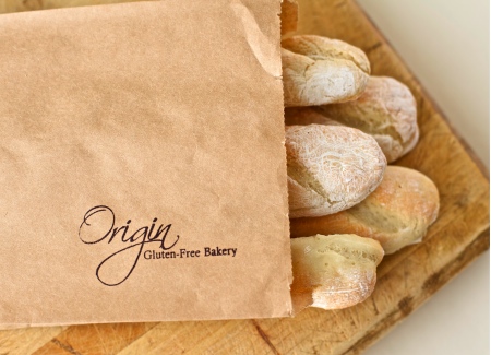 Close up photo of five baguettes poking out of the top of a paper bag with the Origin Gluten Free Bakery logo stamped on the lower right hand corner of the bag, sitting on a wooden breadboard.