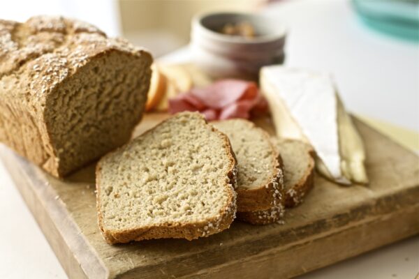 Loaf of bread with three slices on a wooden bread board with a triangle of soft cheese: brie or camembert and charcuterie out of focus in the background