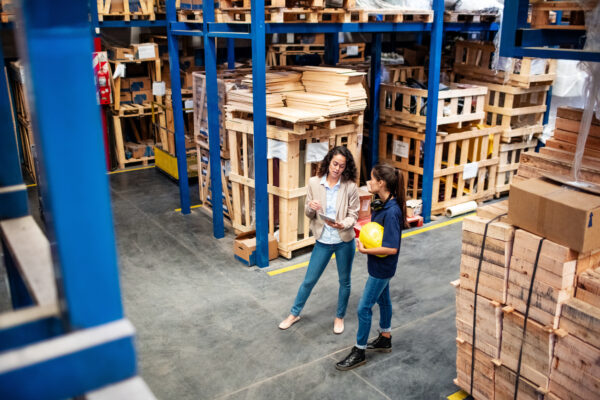 Supervisor holding digital tablet talking with warehouse foreman while standing by a large storage rack. Distribution warehouse employees taking stock inventory.